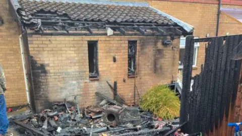 The rear of a house. Its yellow brickwork is charred black and some of the roof is crumbling. There is rubble and debris in the back garden.