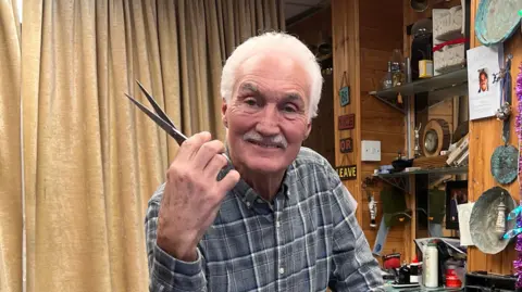 Paul, who has short white hair and a moustache, poses in his salon while holding a pair of scissors. Shelves packed with items can be seen in the background and decorations hanging from the wall.