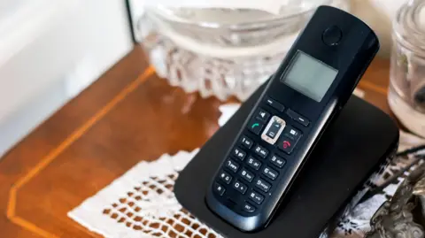 Getty Images A black landline phone, switched off, in its charging stand. The charging stand is on a brown table, which had a white doily on it. There are two glass bowls, one to the left of the phone and on to the right. 