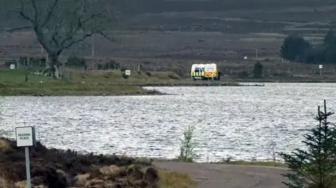 A calm loch with rippling water in the foreground, small evergreen trees along the near shore, and a lone person standing by the water’s edge across the lake. A white van with a yellow panel is parked on the far bank, with open moorland and low hills stretching into the background under an overcast sky.