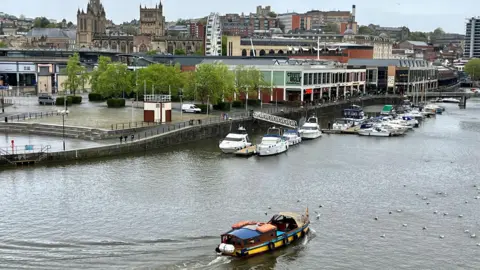 A Bristol Ferry Boat company vessel makes it way across the Floating Harbour towards the city centre. There are many gulls on the water and the city centre is visible in the background
