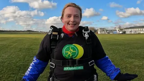 Belinda Dowling Belinda Dowling smiles into the camera with her arms slightly outstretched to her sides. She is wearing a long-sleeved blue top with a black short sleeved top over the top bearing the Air Ambulance logo and her parachute harness goes over her shoulders and around her middle.