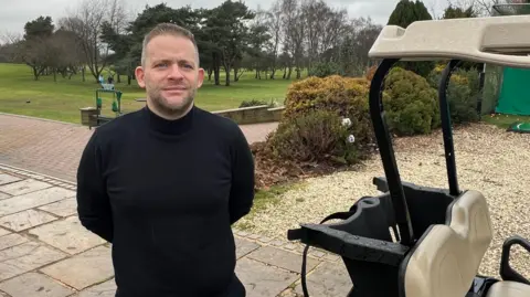 Chris Goodall, general manager at Selby Golf Club, who said repeated power cuts have cost the business thousands of pounds, is standing next to a golf buggy with the course in the background. He is wearing a black jumper and trousers and has short brown hair.