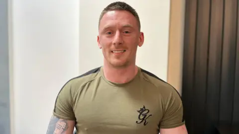 Ben Hellon wears a green T-shirt, standing in the living room of the Own Merit house