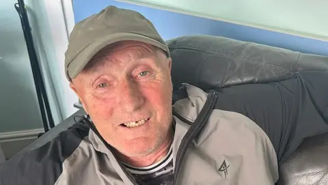 BBC/Melanie James Stuart Hopwood reclining on a black leather sofa, wearing a grey and black jacket and a light-coloured cap. The image is taken from a slightly elevated angle, with a pale blue wall and part of a doorway visible in the background.