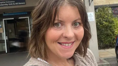 Carly smiles at the camera outside a hospital entrance. She has short brown hair, green eyes and a nose ring. She is wearing an oatmeal brown sweater.