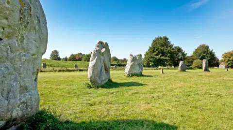 The Avebury stone circle consists of ancient stones placed in a circle on a grass fields with some trees in the background.