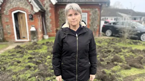 Edd Smith/BBC Caroline Harris, with grey hair and a black jacket, standing by her heavily damaged lawn.