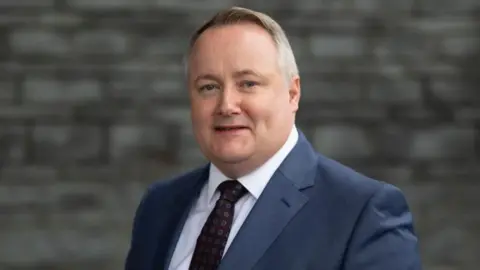 Darren Millar, wearing a blue suit, burgundy and blue polka dot tie and white shirt, in front of the Senedd's slate facade.