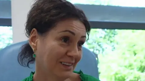 A close-up of Madalina Chifu. She is sitting in a chair in a clinical setting, and is wearing a green top and has short dark hair partially tied back. 
