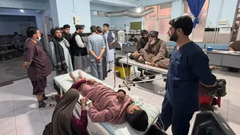 A young male is seen lying down on a stretcher in a hospital ward, hooked up to a drip, as a woman kneels by his side and holds his hand. A man in scrubs is seen stood next to the stretcher looking on at a group on men gathered at the end of the stretcher, while another person can be seen lying on a stretcher in the background