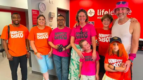 Dementia Disco Three men, two women, one older boy and a younger boy and girl are all smiling while wearing Dementia Disco T-shirts and standing in a reception area as a Life Leisure Centre in Stockport.