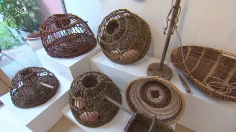 Woven willow crab pots are arranged on white plinths in an art gallery.