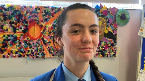 A schoolgirl is standing with art behind her. Her brown hair is in a plait. Her school uniform is blue.