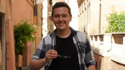 George Webster George Webster is pictured wearing a black and white checked shirt that is open over a black T-shirt. He has short dark hair and is pictured standing outside, holding a pair of glasses in front of his chest. He is smiling directly at the camera. There are honey-coloured stone buildings behind him and some shrubbery. The weather looks sunny.