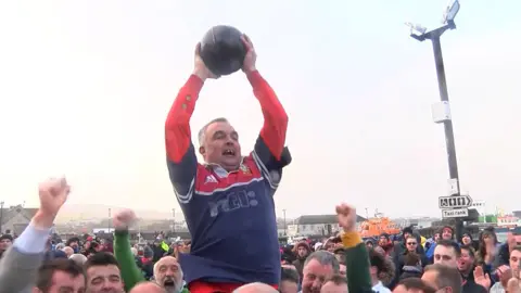 A man jumping above a crowd, he is wearing a red and blue sports top and holding a brown ball.