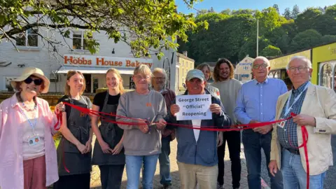 A group of people holding a paper sign with a red ribbon being cut in front of a bakery in Nailsworth