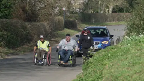 Shaun Whitmore/BBC A woman in black clothing and a baseball cap walking on a country lane beside two men in wheelchairs, one in hi-vis and dark clothing, the other in a grey hoodie and dark trousers. All three are wearing baseball caps. A blue car is following them