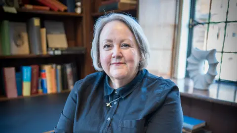 Claire Malcolm, a woman with grey hair and a blue shirt sat in front of a book case.