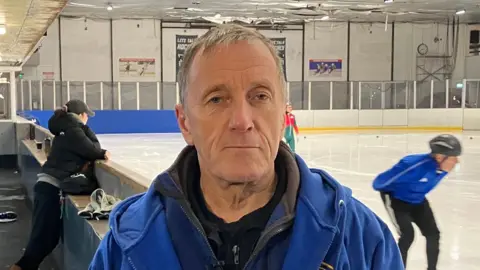 A man with short brown hair stands at the side of an ice rink. He is wearing multiple blue jackets and a black T shirt.