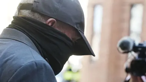 PA Media Robert Bush arrives at Hull Crown Court. He is wearing a dark suit, a dark-grey  baseball cap and a heavy scarf, which covers much of his face below his eyes.