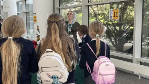 A group of children stood around a woman. The woman is holding a miniature model of a rocket