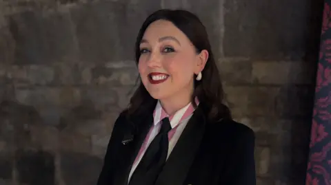 A woman wearing a black suit and red lipstick smiles at the camera 
