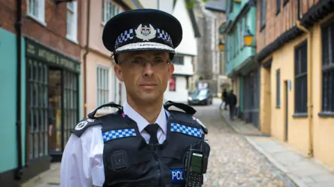 Norfolk Police Chief Constable Paul Sanford standing in uniform on a cobbled street in Norwich. He is wearing glasses and a peaked hat