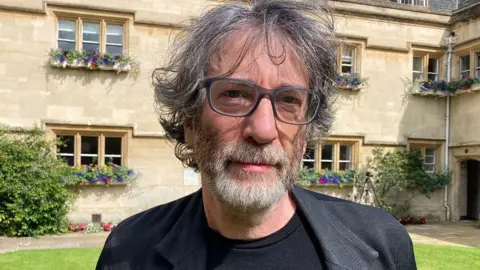 Neil Gaiman pictured outside on the lawn of a property wearing a smart grey jacket and glasses