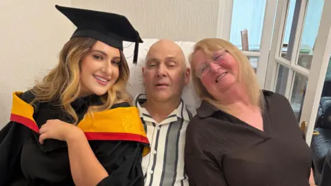 Danielle is wearing a cap and gown and leaning next to her dad in his bed with her mother on his other side. They are smiling at the camera. Her father has a stripy pyjama top on and her mother has thin blonde hair and a brown shirt on. His bed is near a door.