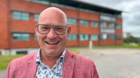 Toby Parkins wearing glasses, a blue and white patterned shirt and a salmon-pink blazer