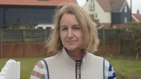 Teresa Lane smiles at the camera from her garden. She has short blonde hair and wears a wool fleece jacket.