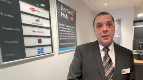 Man in dark suit and striped tie with a Post Office badge stands in front of a board listing the days each of the banks has a representative in the Banking Hub.