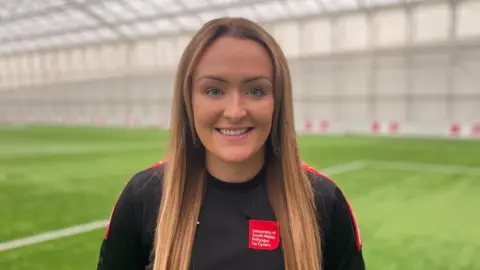 Sara Gray has long brown straight hair. She is wearing a black t-shirt with a University of South Wales red logo below her left shoulder. She is standing on a football pitch.