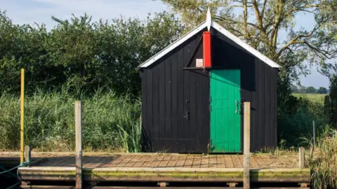 Historic England A small black wooden hut. It has a green door and red sign. In front is a small wooden dock. 