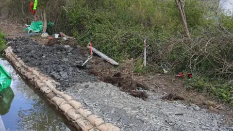 Canal & Rivers Trust Building work is under way by the side of the canal with grey concrete lay on the ground with bricks separating it from the canal. Trees and shrubbery is growing on the right of the repair work.