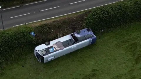 BBC A bus overturned on grass near a main road 