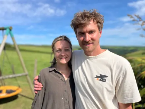 A man in his mid thirties with a beard and tousled hair has his arm around a woman in her mid thirties. She has a dark grey blouse on and they're standing in a garden in front of children's playing equipment on a bright summer's day.