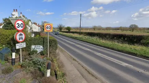 A road in a rural area with road signs to the left, one saying 'Gate Helmsley'
