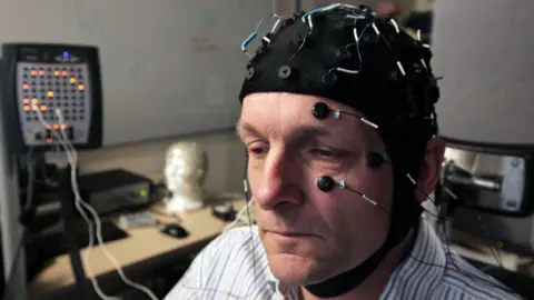 Michael Mosley wearing a special cap and numerous sensors attached to his head and face, attached to a medical machine