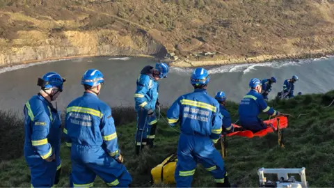 A coastguard team stood on the edge of a grassy cliff above water with rescue equipment