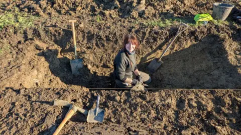 Woman sat inside a ditch dug into earth at Historic England's excavation site, near Willersey. She is wearing black jumper and trousers and has red hair. 
