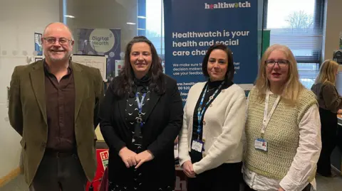 One man and three women stand in a line. They are the health hub team. They are standing in front of a Healthwatch banner.