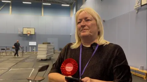 A woman is standing in a sports hall after an election. She is wearing a dark top and a red rosette.