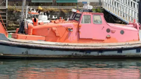 Jemma Sworder An orange lifeboat, in poor condition with chipped and faded paint, is docked in a harbour.