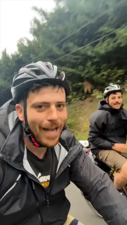 Two men on bikes. They are wearing coats and helmets. There are trees in the background. 