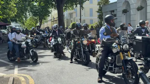 A large group of men on motorbikes in a town centre on a sunny day as part of the Distinguished Gentleman’s Bike Ride. They are all wearing smart shirts, waistcoats or a full three piece suits. 