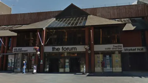 A general view of entrance to The Forum in Barrow, Cumbria. The signs above the glass doors and windows are black and white. There is a triangular design on the roof above the main door. There is a man walking across the front of the building, and flowers in the hanging baskets.