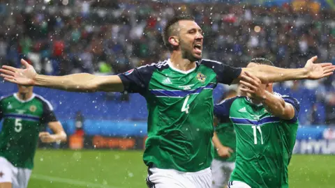Getty Images Gareth McAuley (L) of Northern Ireland, in a green shirt with blue trim, celebrates scoring his team's first goal with his team mate Conor Washington (R) during the UEFA EURO 2016 Group C match between Ukraine and Northern Ireland at Stade des Lumieres on June 16, 2016 in Lyon, France.
He has his arms outstretched and mouth open as hailstones fall around them.