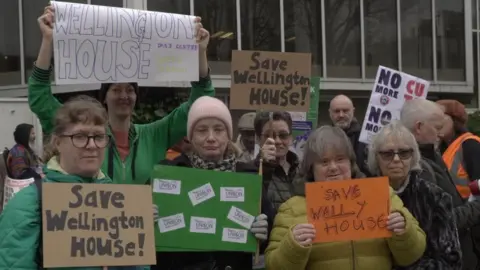 LDRS/Oliver Leader de Saxe A group of people holding placards and posters, which read "save Wellington House". They are standing outside a local council building.